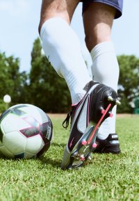 Black and silver soccer cleats with pink accents, partially on grass, next to a multi-colored soccer ball. White knee-high socks visible.