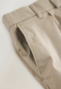 Beige Baumwollhosen mit flachem Bund, diagonalen Taschen und glatter Textur, die über saubere Nähte entlang der Kanten verfügen.