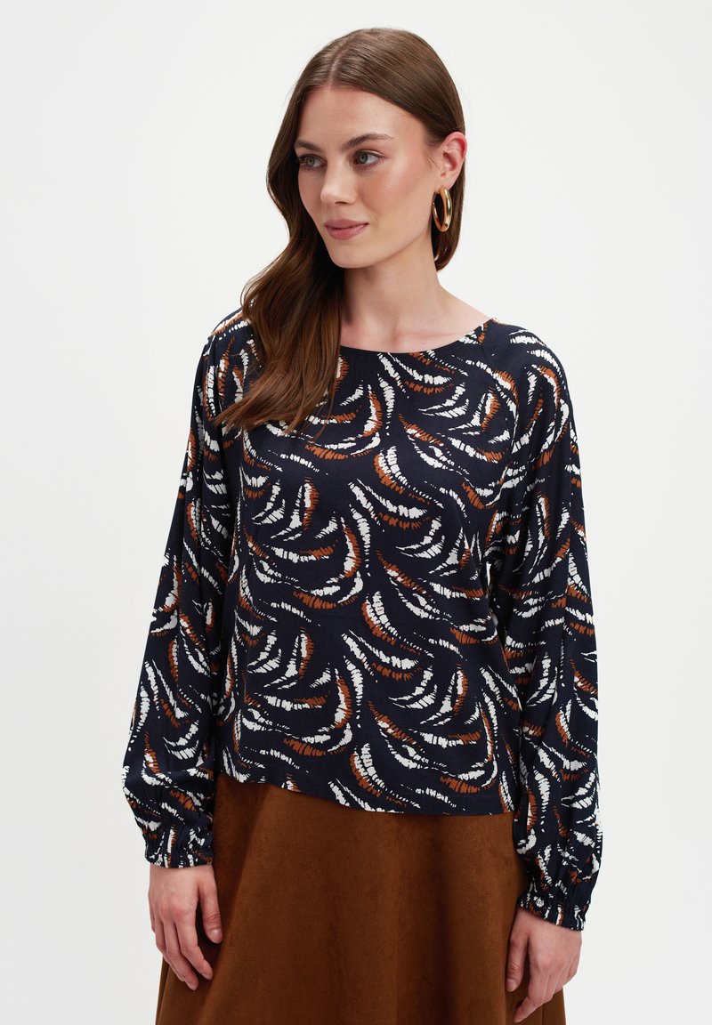 Kobieta z długimi brązowymi włosami, nosząca złote kolczyki w kształcie koła oraz granatową bluzkę z białymi i brązowymi abstrakcyjnymi wzorami piór, w połączeniu z brązową spódnicą.