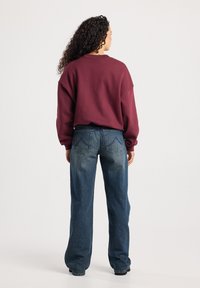 Vrouw met krullend haar staat met de rug naar de camera, gekleed in een bordeauxrode sweatshirt en hoge blauwe jeans met achterzakken.