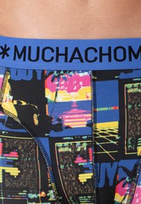 Vīriešu bokseršorti ar zilu elastīgu jostasvīru, kas atzīmēts ar "*MUCHACHOM*", un krāsainu grafisko apdruku, kas satur neona rakstus uz melna fona.