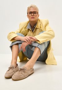 Mocassins en daim beige clair avec lacets, bout arrondi et profil bas. Portés avec un jean bleu retroussé à la cheville et un blazer jaune.