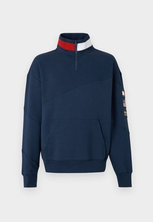 Marineblauer Sweatshirt mit Viertelreißverschluss, roten und weißen Streifen am Kragen, Fronttasche und Aufnähern am linken Ärmel.