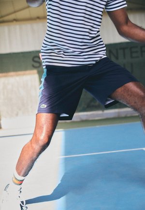 Mandlig tennisspiller i stribet skjorte og marineblå shorts, der dykker på banen, holder en ketsjer, iført hvide sneakers og stribede sokker.