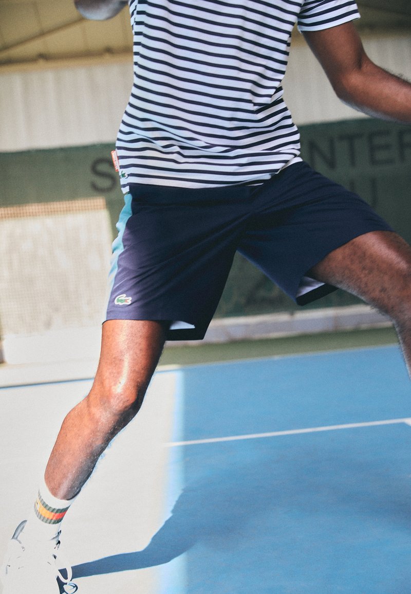 Joueur de tennis masculin en chemise rayée et shorts navy, se jetant sur le court, tenant une raquette, portant des baskets blanches et des chaussettes rayées.