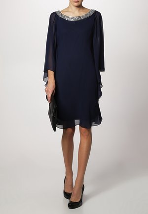 Robe bleu marine avec des manches en cape transparentes, un décolleté orné et une silhouette fluide, assortie à une pochette noire et des talons hauts.