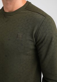 Felpa verde oliva con un motivo testurizzato, collo tondo e una piccola etichetta del brand sul petto. Realizzata in un tessuto morbido ed elastico.