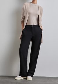 Černé kalhoty na míru s hladkou texturou, doplněné světle béžovým topem a sladěným dlouhým kardiganem, nošené s bílými teniskami.
