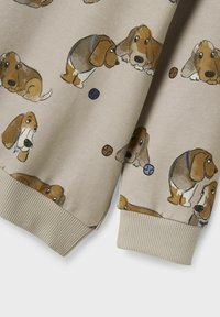 Beige sweatshirt met herhaalde cartoonontwerpen van een basset hound, met blauwe en bruine ballen. Geribbelde manchetten en onderkant zorgen voor een tekstuurlijke afwerking.