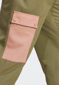 Olive green pants with a pink fabric pocket featuring snap closures. Smooth texture, tapered design, and simple construction details.