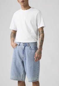 Mężczyzna ubrany w gładki biały t-shirt z krótkim rękawem i jasnoniebieskie denimowe szorty, z tatuażami na ramionach i jedną ręką w kieszeni.