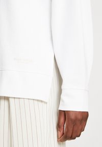 Felpa bianca con vestibilità comoda, polsini e orlo a coste. Presenta un marchio discreto vicino all'orlo. Abbinata a pantaloni beige a righe.