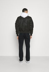 Giubbotto bomber nero in tessuto testurizzato, con polsini a coste e una felpa con cappuccio bianca sotto, abbinato a jeans scuri e sneakers nere.