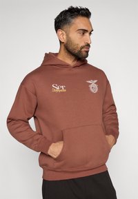 Brązowy hoodie wykonany z miękkiego materiału z przednią kieszenią, zdobiony białymi nadrukami, w tym herbem i słowem "Ser" w złotym kolorze.