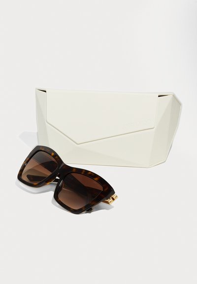 Lunettes de soleil écaille de tortue avec des verres sombres et des détails en or, placées à côté d'une coque géométrique blanche avec un logo délicatement embossé.