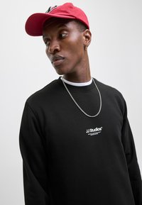 Sudadera negra con logotipo blanco en el pecho, combinada con una gorra roja y un collar de cadena plateado; textura suave y corte relajado.