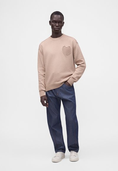 Mannequin masculin portant un pull beige avec un cœur texturé sur la poitrine, jean bleu et baskets blanches, debout avec une main dans la poche.