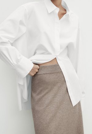 Person wearing an oversized white button-up shirt partially tucked into a high-waisted beige knit skirt, hand resting near waistband.