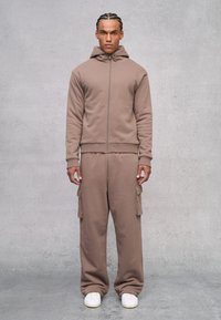 Bruine zip-up hoodie met een fitted zoom en een capuchon. Gekoppeld aan bijpassende losse cargo broek met zijzakken en witte sneakers.