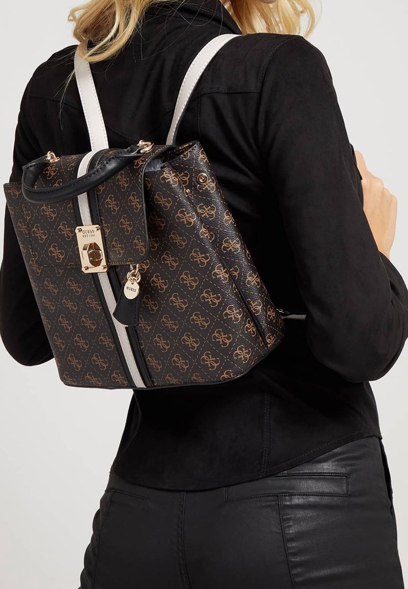 Femme portant une veste et un pantalon noirs avec un sac à dos Guess marron à motifs, doté de bretelles blanches et de détails métalliques dorés.