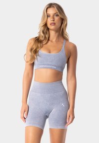 Gaiši pelēka sporta krūšturis un augstas jostas šorti, kas izgatavoti no mīksta auduma, ar mazu logo uz šortiem un joslu ar zīmola nosaukumu.