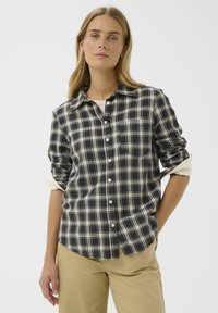 Donna con lunghi capelli biondi che indossa una camicia a quadri bianca e nera con maniche arrotolate e pantaloni beige, in piedi con una mano in tasca.