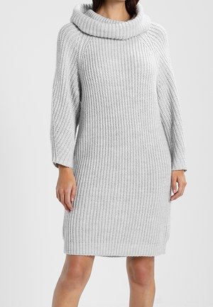 Kobieta w jasnoszarej, prążkowanej dzianinowej sukience z długimi rękawami i szerokim, złożonym golfem.