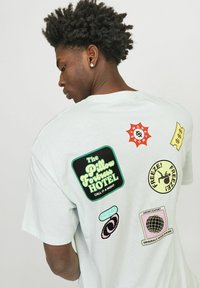 Camiseta de algodón verde menta con varios parches gráficos en colores brillantes, incluyendo un logo de hotel y divertidos íconos en la parte de atrás.