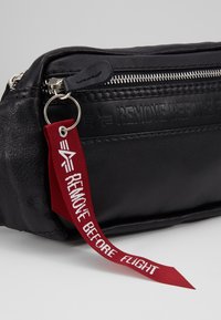 Musta nahkainen pussukka, jossa on vetoketjusulku, korotetulla tekstimuotoilulla ja punaisella kangastarralla, jossa lukee "POISTA ENNEN LENTOA."