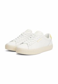 Baskets en cuir blanc avec semelle en caoutchouc texturé, présentant un accent jaune sur le talon et les lacets, et un design minimaliste.