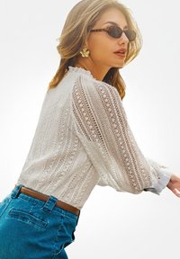 Donna che indossa occhiali da sole tartarugati, grandi orecchini a forma di fiore, una blusa di pizzo bianco e jeans blu con una cintura marrone, di profilo.