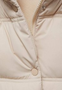 Beige vadderad jacka med en mjuk yta, som har en dragkedja och tryckknappsstängning. Utmärkande quiltad design längs kroppen.