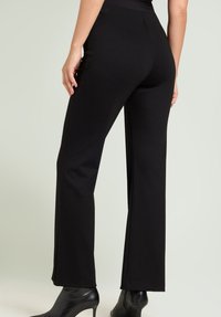 Zwarte, high-waisted broek gemaakt van rekbare stof met een rechte pijp, gedetailleerd met minimale naden en gedragen met zwarte hakken.