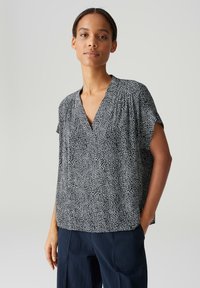 Blusa bianca e nera con scollo a V, maniche corte e un motivo testurizzato che sembra piccole forme irregolari. Vestibilità ampia, design casual.