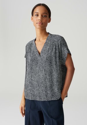 Schwarz-weiße Bluse mit V-Ausschnitt, kurzen Ärmeln und einem strukturierten Muster, das kleinen unregelmäßigen Formen ähnelt. Lockere Passform, lässiges Design.