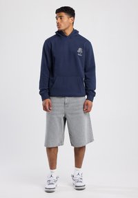Sudadera navy con un bolsillo frontal y detalle de logo, combinada con pantalones cortos de mezclilla grises. Los zapatos son blancos con detalles en negro.