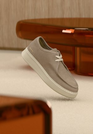 Lichtegrijze suède casual schoen met witte zool en veters zwevend boven een beige tapijt in een kamer met amberkleurige meubels.