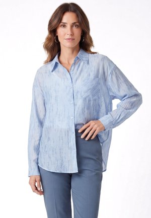Frau trägt hellblau gemustertes Hemd mit Knöpfen und schieferblaue Hose, steht mit einer Hand in der Hüfte vor weißem Hintergrund.