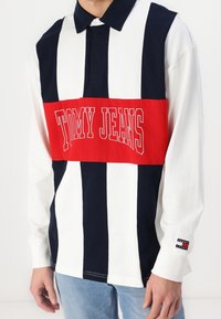 Long-sleeve polo shirt in navy, white, and red stripes. Features a buttoned collar, central red panel with "TOMMY JEANS" text, and branded cuff.
