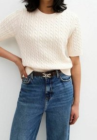 Nainen, jolla on päällään kerma beige kaapelikudottu lyhythihainen neulepaita, joka on pujotettu korkeavyötäröisiin sinisiin farkkuihin. Vyötä koristi musta vyö, jossa on kultainen puolikuu-solki.