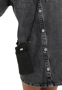 Bolso cruzado de cuero negro con un cierre superior con cremallera, que presenta una etiqueta de marca en tono dorado. Combinado con un vestido camisa de denim gris.