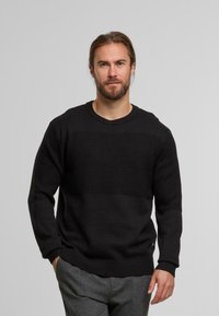 Schwarzer Strickpullover mit strukturiertem Oberteil, gerippten Ärmelbündchen und Saum. Getragen mit grauen Hosen; das Material wirkt weich und dick.
