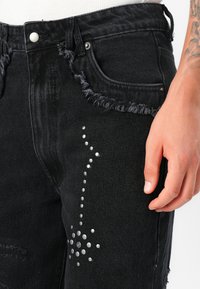 Vaqueros de mezclilla negros con cintura y bolsillos deshilachados. Presenta clavos plateados dispuestos en un patrón de puntos en el muslo delantero.