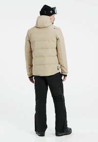 Beige Pufferjacke mit Kapuze und gestepptem Design, getragen mit schwarzen Hosen und Stiefeln. Verfügt über ein Logo an der Schulter und Reißverschlusstaschen.