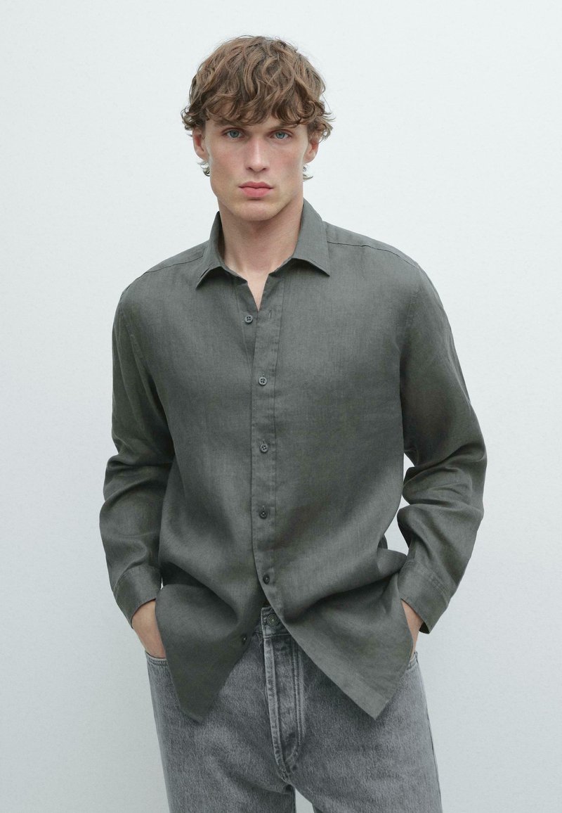 Jeune homme aux cheveux bouclés portant une chemise grise ample à boutons et un jean gris clair, debout les mains dans les poches devant un fond uni.