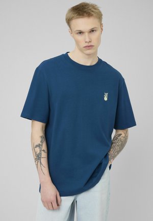 Jeune homme aux cheveux blonds courts portant un T-shirt bleu foncé uni avec un petit emblème clair et un jean bleu clair, montrant des tatouages sur le bras.