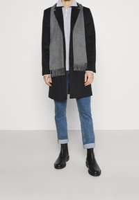 Schwarzer Mantel über einem grauen Reißverschluss-Pullover, kombiniert mit hochgekrempelten blauen Jeans und schwarzen Stiefeletten. Ein grauer Schal mit Fransen sorgt für zusätzliche Textur.