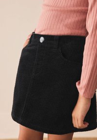 Gonna in velluto a coste nera con superficie testurizzata, caratterizzata da chiusura con bottoni e tasche laterali, indossata con una maglia a costine rosa a maniche lunghe.