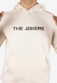 Sudadera con capucha color crema que incluye un bolsillo frontal, una capucha con cordón y el texto "THE JOKERS" en negro, con un acabado texturizado o estampado.