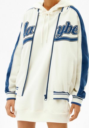 Persoon die een witte hoodie-jurk draagt en een wit-blauwe varsityjas met gestreepte manchetten en letters over de borst.